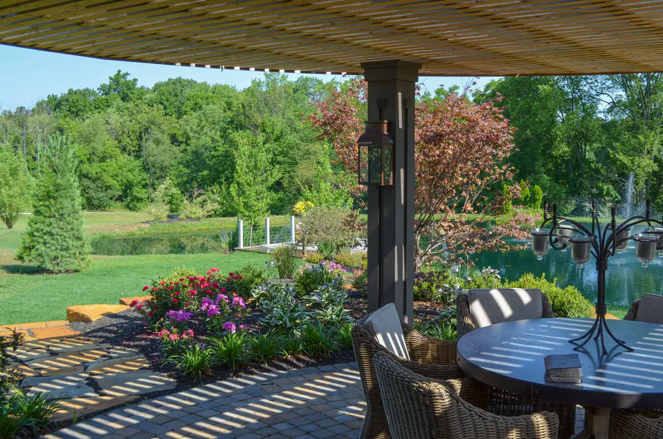Dining table under covered patio with paver floor, wood ceiling, columns, planting beds, and long view across the property.